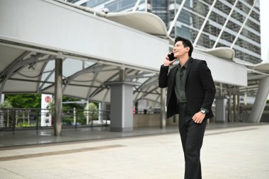 Takım elbiseli bir adam dışarıda dururken cep telefonuyla konuşuyor. Keyfi yerinde ve profesyonel olarak giyinmiş...