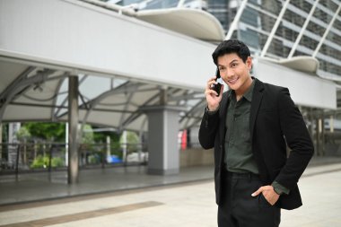 Takım elbiseli bir adam dışarıda dururken cep telefonuyla konuşuyor. Keyfi yerinde ve profesyonel olarak giyinmiş...