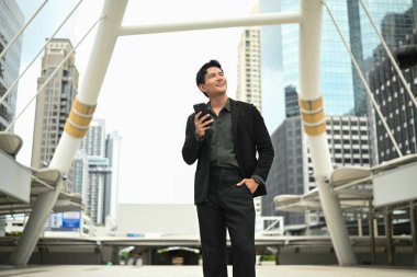 Takım elbiseli bir adam yüksek bir binanın önünde duruyor, elinde cep telefonu var...