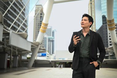 Takım elbiseli bir adam yüksek bir binanın önünde duruyor, elinde cep telefonu var..