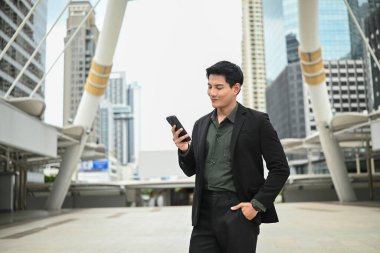 Takım elbiseli bir adam yüksek bir binanın önünde duruyor, elinde cep telefonu var...