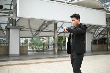 Takım elbiseli ve kravatlı bir adam saatine bakıyor. Bir iş ortamında ve acelesi var..