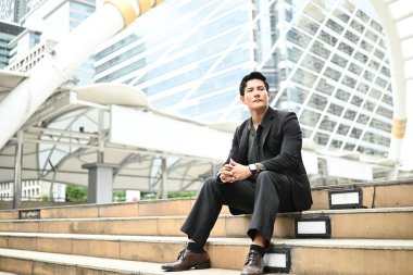 Takım elbiseli bir adam büyük bir binanın önündeki basamaklarda oturur. Bir saat takıyor ve birini bekliyor..