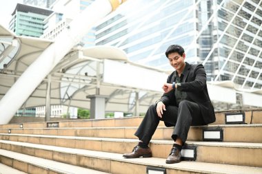 Takım elbiseli ve kravatlı bir adam merdivenlerde oturuyor. Saat takıyor ve saati kontrol ediyor..