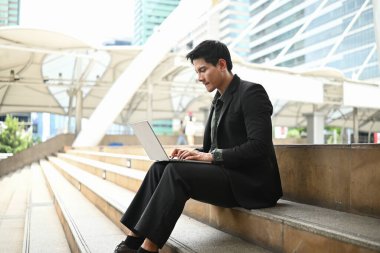 Takım elbiseli bir adam önünde dizüstü bilgisayarla merdivenlerde oturuyor. Ekrana odaklandığı için çalışıyor ya da çalışıyor...