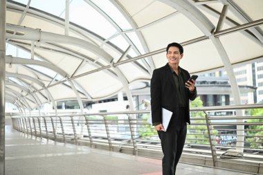 Takım elbiseli bir adam elinde cep telefonu ve diğer elinde dizüstü bilgisayarla yürüyor. Profesyonellik ve meşguliyet kavramı.