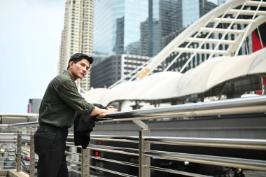 Yeşil gömlekli ve siyah ceketli bir adam şehrin siluetinin önündeki köprüde duruyor...