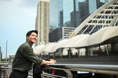 Bir adam gülümsüyor ve büyük bir binanın önündeki korkuluklara yaslanıyor. Adam şehrin manzarasının tadını çıkarıyor.