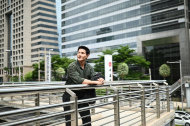 Bir adam gülümsüyor ve büyük bir binanın önündeki korkuluklara yaslanıyor. Adam şehrin manzarasının tadını çıkarıyor..