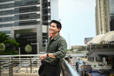 Bir adam gülümsüyor ve büyük bir binanın önündeki korkuluklara yaslanıyor. Adam şehrin manzarasının tadını çıkarıyor..