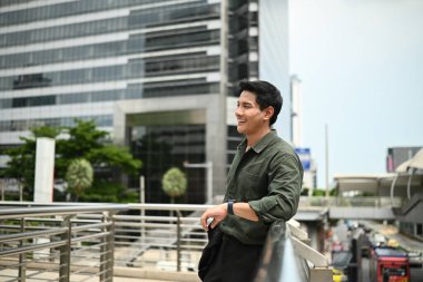 Bir adam gülümsüyor ve büyük bir binanın önündeki korkuluklara yaslanıyor. Adam şehrin manzarasının tadını çıkarıyor..