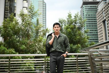 Yeşil gömlekli ve siyah ceketli bir adam şehrin siluetinin önündeki köprüde duruyor...