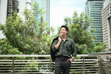 Yeşil gömlekli ve siyah ceketli bir adam şehrin siluetinin önündeki köprüde duruyor...