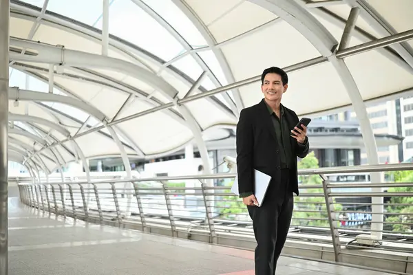 Takım elbiseli bir adam elinde cep telefonu ve diğer elinde dizüstü bilgisayarla yürüyor. Profesyonellik ve meşguliyet kavramı.