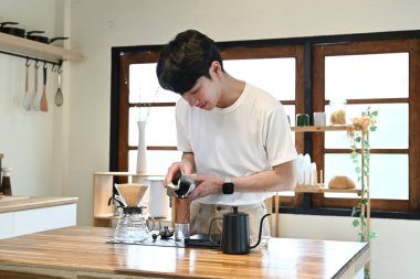 Bir adam mutfakta kahve yapıyor. O bir fincana kahve dolduruyor ve beyaz bir gömlek giyiyor..