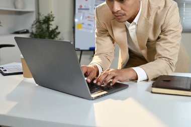 Takım elbiseli bir adam önünde bir dizüstü bilgisayarla masada oturuyor. Klavyeye yazı yazıyor ve işine odaklanmış durumda...