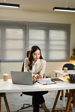 Takım elbiseli bir kadın bir masada dizüstü bilgisayarı ve bir yığın kağıtla oturuyor. İşine odaklanmış durumda.