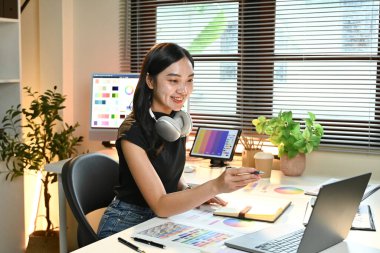 Masasında dizüstü bilgisayarı ve not defteri olan bir kadın oturuyor. Gülümsüyor ve mutlu. Masa bir saksı bitkisi, bir klavye ve bir fare gibi çeşitli eşyalarla dolu.