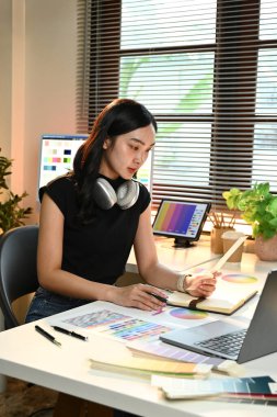 Masasında dizüstü bilgisayarı ve not defteri olan bir kadın oturuyor. Bir proje üzerinde çalışıyor ve odaklanmış durumda.