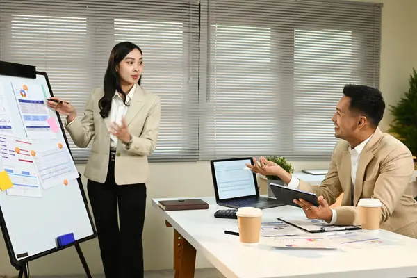 Bir kadın takım elbiseli bir adama sunum yapıyor. Adamın elinde bir tablet var ve kadın beyaz tahtayı işaret ediyor.