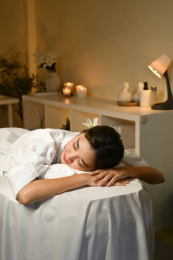 Beyaz bornozlu bir kadın masaj masasında yatıyor. Gülümsüyor ve rahatladı.
