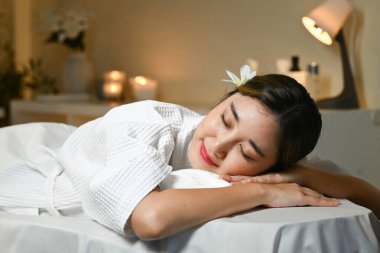 Beyaz bornozlu bir kadın masaj masasında yatıyor. Gülümsüyor ve rahatladı..