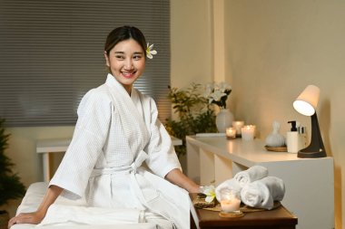 Bir kadın spa 'da bir yatakta oturuyor, beyaz bir bornoz giyiyor ve gülümsüyor. Oda çiçekler ve mumlarla süslenmiş, rahatlatıcı bir atmosfer yaratıyor..