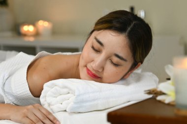 Beyaz bornozlu bir kadın masaj masasında yatıyor. Gülümsüyor ve rahatladı..