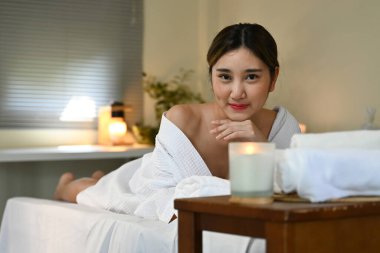 Beyaz bir bornoz ve önünde mum olan bir kadın spa yatağında yatıyor. Gülümsüyor ve eğleniyor...