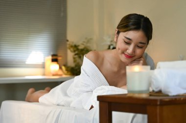 Bir kadın masaj masasında kaplıcada yatıyor. O gülümsüyor ve beyaz bir bornoz giyiyor. Oda loş ve rahatlatıcı bir atmosferi var..