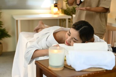 Sırtında bir şişe losyonla spa yatağında yatan bir kadın. Arkasında duran bir kadın losyonu sırtına döküyor. Rahatlama ve bakım kavramı.