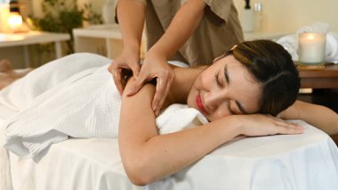 Bir kadın masaj terapistinden masaj alıyor. Gülümsüyor ve rahatlıyor. Masaj masası beyaz bir havluyla kaplı.