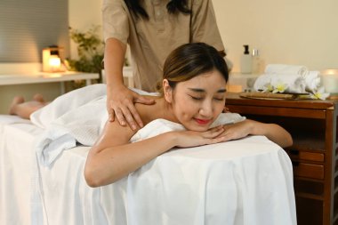 Bir kadın masaj terapistinden masaj yaptırıyor. Kadın gülümsüyor ve masajın tadını çıkarıyor. Oda loş bir şekilde aydınlatılmış ve yakınlardaki bir masada mumlar var..