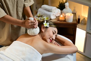 Sıcak bitkisel spa masajı alan bayan, masör masaj salonundaki mumların sıcacık ışığında kadına nazikçe şifalı bitki torbalarını bastırıyor..