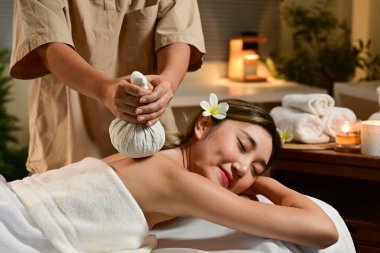 Sıcak bitkisel spa masajı alan bayan, masör masaj salonundaki mumların sıcacık ışığında kadına nazikçe şifalı bitki torbalarını bastırıyor..