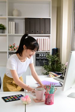 Young asian woman is stand at a desk with a computer in office . She is smiling and she is happy.