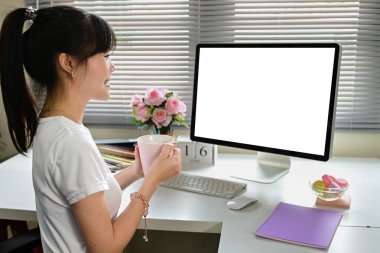 Bir kadın bilgisayar monitörü ve bir fincan kahveyle masada oturuyor. Gülümsüyor ve işinden zevk alıyor. Masada klavye, fare ve bir kase şeker var.