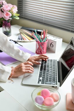 Beyaz bir masanın önünde dizüstü bilgisayarda yazan bir kadın. Masada bir kase pembe badem kurabiyesi, çiçekli bir vazo, bir fincan, bir kitap gibi çeşitli eşyalar var.
