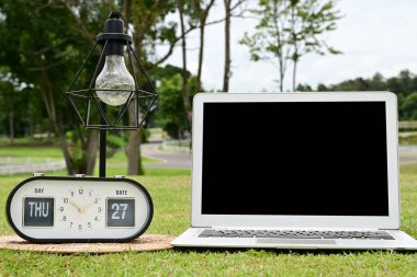 Siyah ekranlı beyaz bir dizüstü bilgisayar yeşil çimlerin üzerine yerleştiriliyor, yanında bir lamba ve bir saat var...