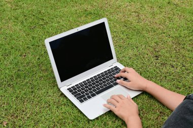 Bir kişi çimenli bir alanda dizüstü bilgisayarda yazıyor. Laptop açık ve ekran siyah..