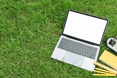 Bir dizüstü bilgisayar çimenli bir alanda, bir saat ve bir not defteriyle birlikte. Laptop açık ve çimenler yeşil.