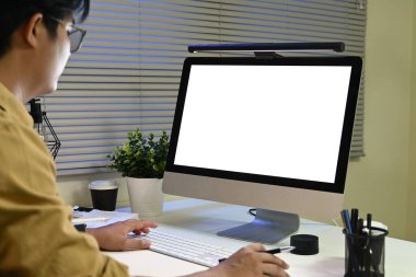 Beyaz arkaplanı olan bir kişi bilgisayar monitörüne yazı yazıyor. Gözlük takıyor ve önünde saksı bitkisi olan bir masada oturuyor...