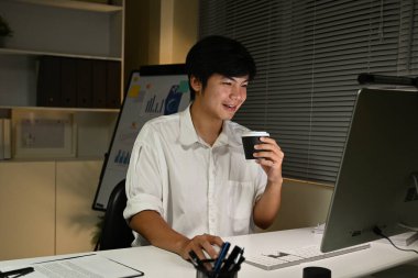 Bir adam bilgisayar monitörü ve bir fincan kahveyle masada oturuyor. Gülümsüyor ve çalışırken kahvesinin tadını çıkarıyor..