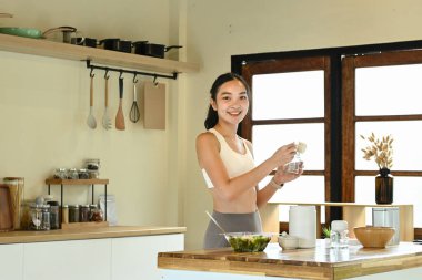 Mutfakta elinde şişeyle gülümseyen bir kadın var. Mutfak bir kase, bir kaşık, bir bıçak ve bir şişe de dahil olmak üzere çeşitli alet ve gereçlerle dolu.