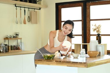 Bir kadın mutfakta, gülümsüyor ve elinde bir konteynır tutuyor. Mutfak bir kase, bir kaşık, bir bıçak ve bir şişe de dahil olmak üzere çeşitli alet ve gereçlerle dolu.