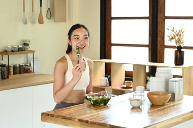 Bir kadın mutfakta salata yiyor. Mutfak çeşitli mutfak aletleri ve kaselerle donatılmıştır..