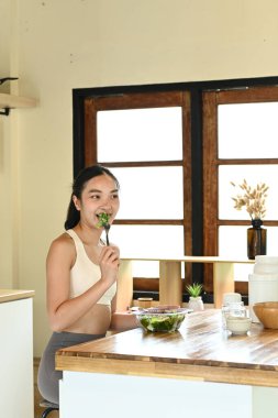 Bir kadın mutfakta salata yiyor. Mutfak çeşitli mutfak aletleri ve kaselerle donatılmıştır..