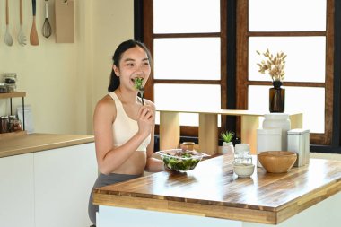 Bir kadın mutfakta salata yiyor. Mutfak çeşitli mutfak aletleri ve kaselerle donatılmıştır..