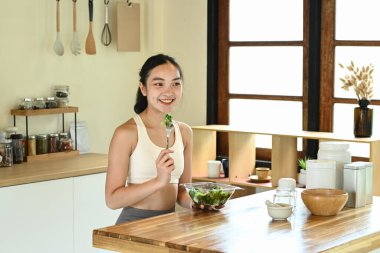 Bir kadın gülümsüyor ve önünde salatayla çatal tutuyor. Masa kâseler, fincanlar ve kaşıklarla dolu. Masanın üzerinde birkaç şişe ve kavanoz var..