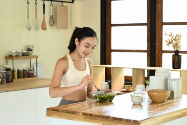 Bir kadın mutfakta salata yiyor. Mutfak çeşitli mutfak aletleri ve kaselerle donatılmıştır.